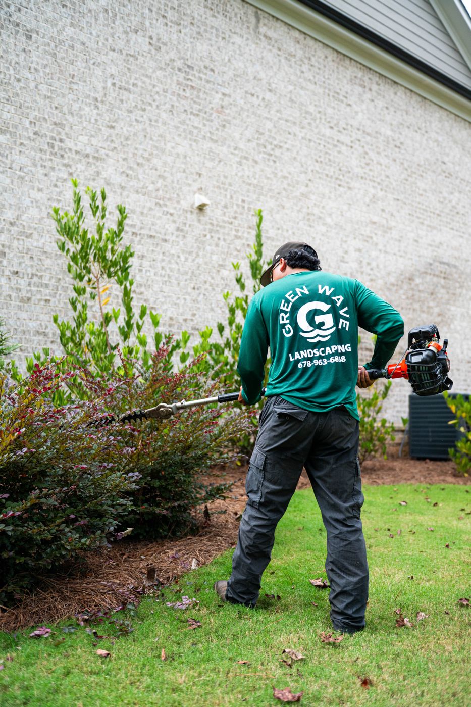 Green Wave crew member trimming shrubs beside a brick home